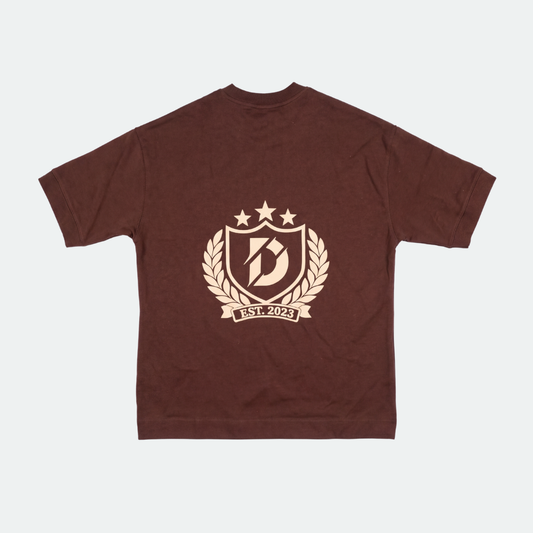 A dark brown short-sleeve t-shirt with a beige graphic print on the front. The graphic features a shield with a stylized letter 'B', three stars, a laurel wreath, and a banner reading 'EST.