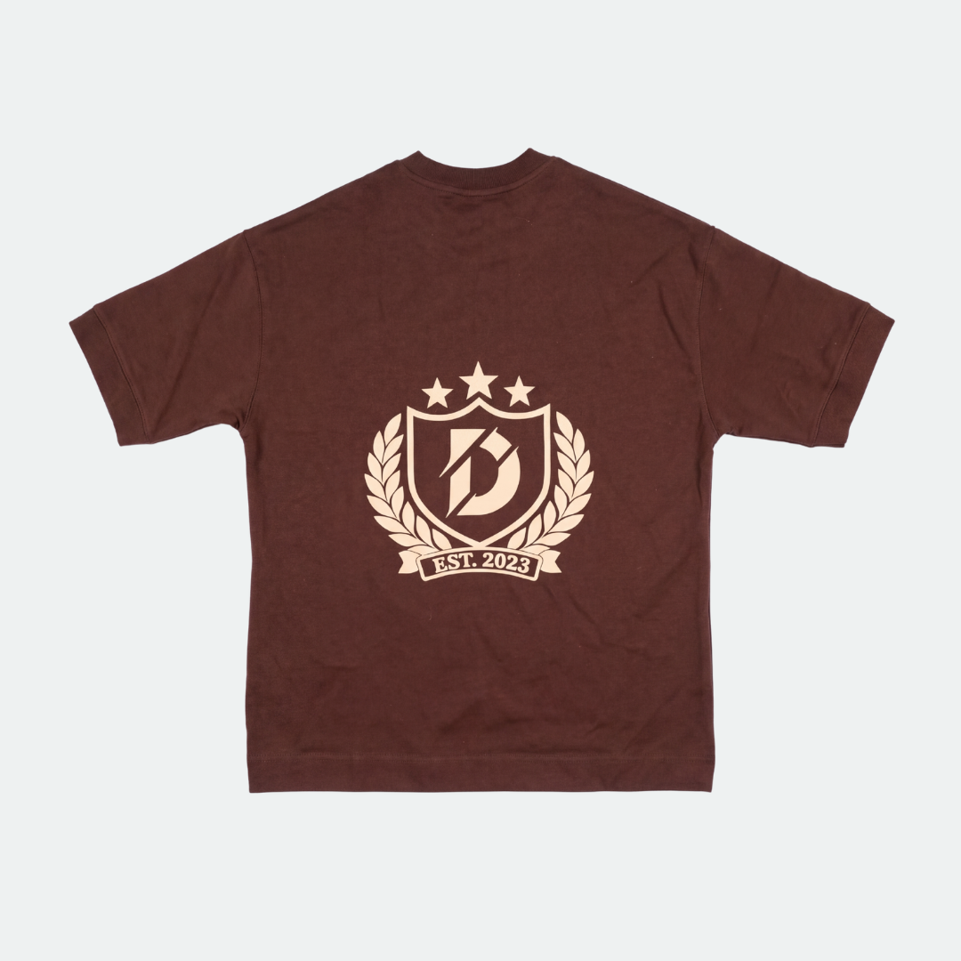 A dark brown short-sleeve t-shirt with a beige graphic print on the front. The graphic features a shield with a stylized letter 'B', three stars, a laurel wreath, and a banner reading 'EST.