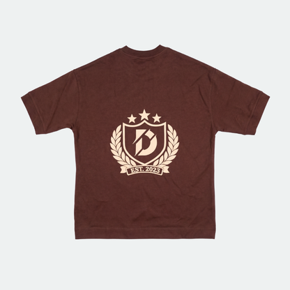 A dark brown short-sleeve t-shirt with a beige graphic print on the front. The graphic features a shield with a stylized letter 'B', three stars, a laurel wreath, and a banner reading 'EST.
